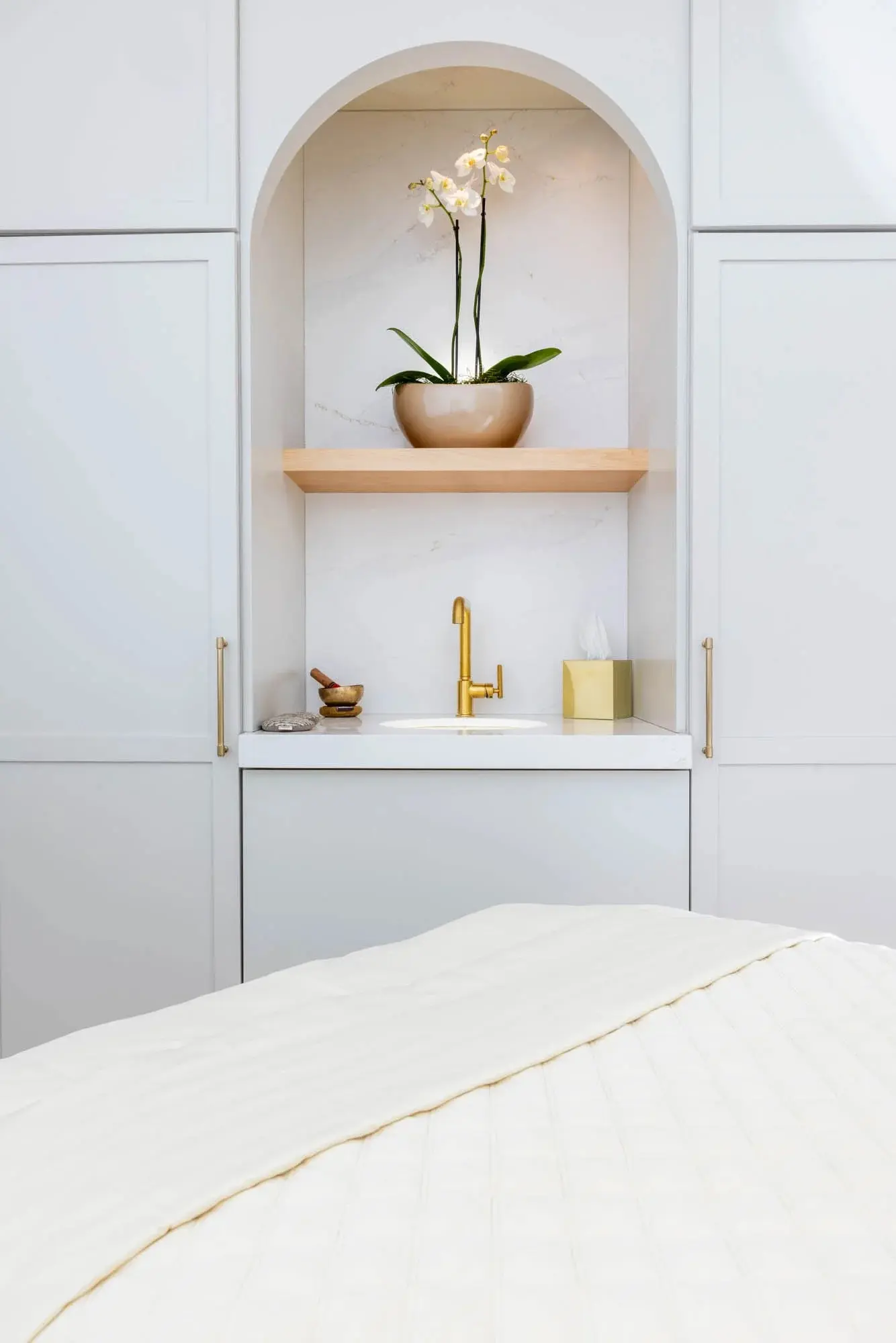 An arched niche with an orchid and brass fixtures beside a made-up treatment table